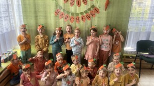 na tle zielonej zasłony z napisem dzień marchewki dzieci pozują do zdjęcia w opaskach z marchewkami na głowie i z marchewkami w ręku